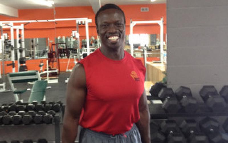 Stephen Menya standing with barbells among free weights. 