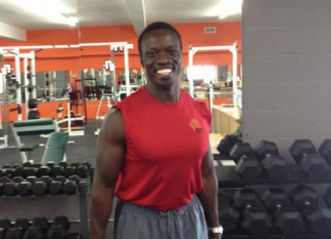 Stephen Menya standing with barbells among free weights. 